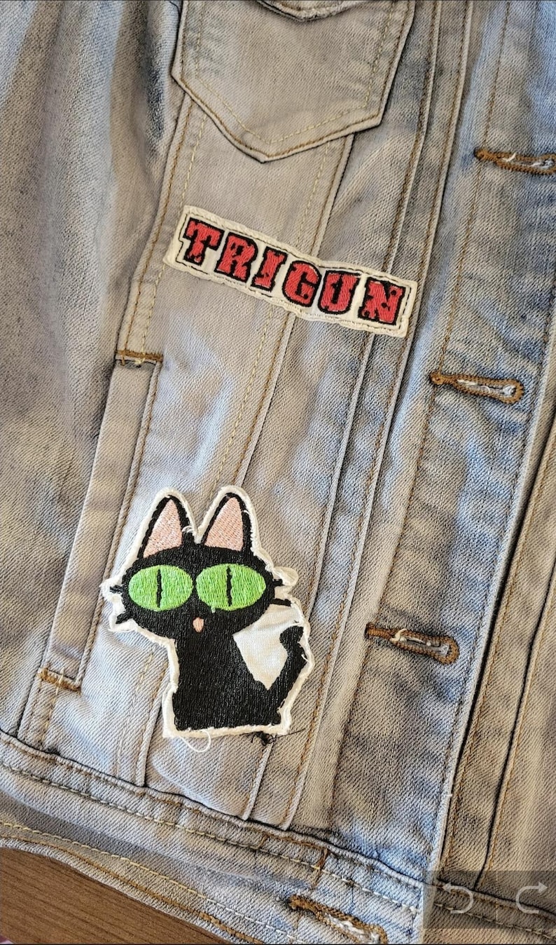 K&ouml;nnte beinhalten: Hellblaue Jeansjacke mit dem Wort "TRIGUN" in roter Schrift auf einem Aufn&auml;her. Ein schwarzer Katzenaufn&auml;her mit gr&uuml;nen Augen und wei&szlig;em Bauch ist ebenfalls sichtbar. Die Jacke hat Metallkn&ouml;pfe und ein klassisches Design.