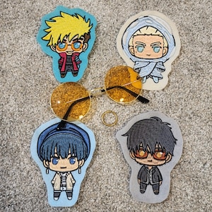 Puede incluir: Cuatro parches de hierro bordados con personajes de estilo anime. Los personajes llevan todos atuendos diferentes y tienen diferentes colores de cabello. Los parches están sobre un fondo gris claro.