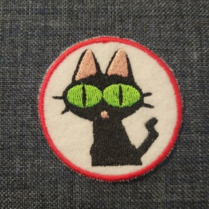 May include: A black cat with green eyes and pink ears is embroidered on a white circle with a red border.
