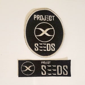 May include: Two black embroidered patches with white stitching. The larger patch is round and says "PROJECT SEEDS" with an infinity symbol in the center. The smaller patch is rectangular and says "PROJECT SEEDS" with an infinity symbol on the left.