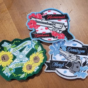 May include: Three embroidered patches featuring different guns and floral designs. The first patch is green with sunflowers and the text "Stangun" and "Chry" on a white banner. The second patch is white with red flowers and the text "Humanoid" and "Typhoon" on a blue banner. The third patch is white with blue flowers and the text "Derriinger" and "Meryl" on a blue banner.