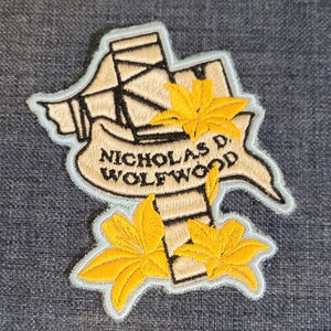 May include: An embroidered patch featuring a stylized image of a gun, two yellow flowers, and a banner with the text "NICHOLAS D. WOLFWOOD".