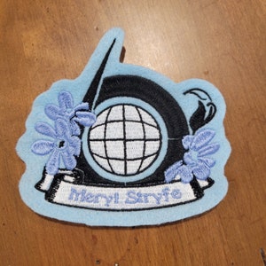 May include: Embroidered patch with a light blue felt backing. The design features a black diving helmet with a white globe, a black sword, and light blue floral accents. The text "Meryl Stryfe" is embroidered on a white banner.