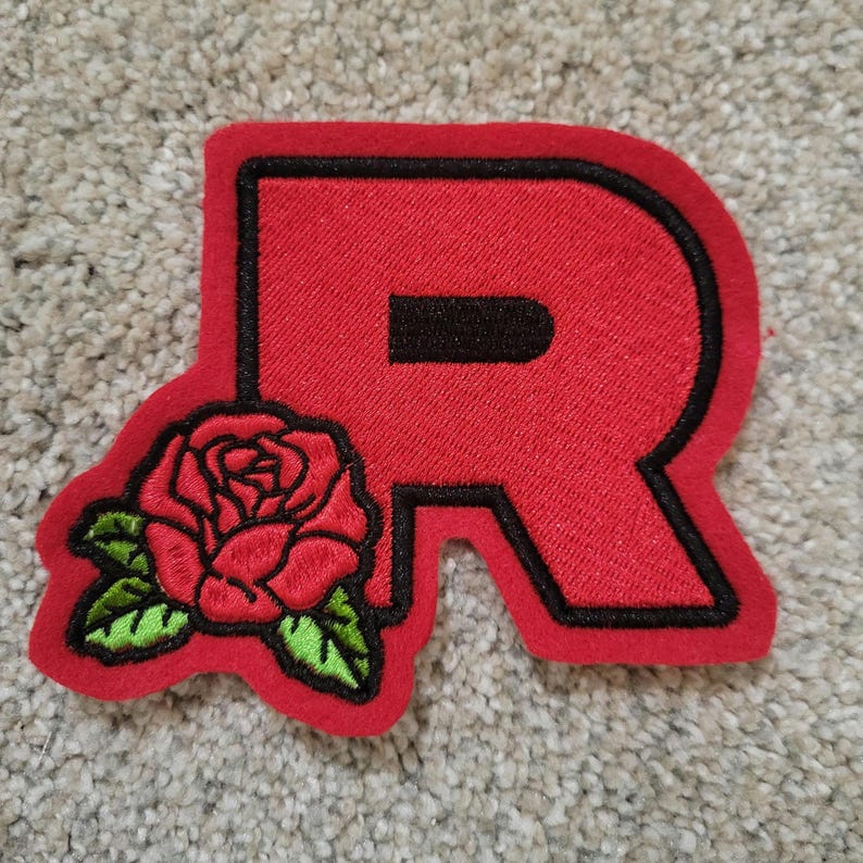 May include: A red felt patch with a black outline of the letter R and a red embroidered rose with green leaves.