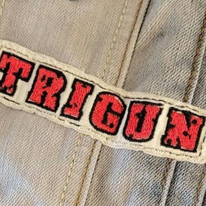 May include: Embroidered patch with the word "TRIGUN" in red letters with a black outline on a white background. The patch is sewn onto a light blue denim jacket.