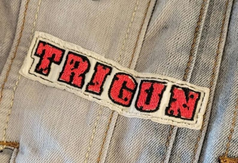 K&ouml;nnte beinhalten: Gestickter Aufn&auml;her mit dem Wort "TRIGUN" in roten Buchstaben mit schwarzer Umrandung auf wei&szlig;em Grund. Der Aufn&auml;her ist auf eine hellblaue Jeansjacke gen&auml;ht.