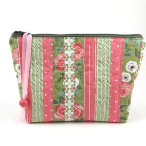 Peut inclure: Une trousse de maquillage matelassée avec une fermeture éclair verte et un ruban rose. Le sac présente des rayures verticales de tissu rose, vert floral et blanc à motifs. Le motif comprend des roses et des cœurs.