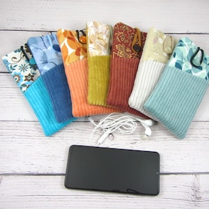 Peut inclure: Plusieurs étuis de téléphone en tissu colorés avec divers motifs et textures, dont des motifs floraux, en velours côtelé et à motifs. Un smartphone noir et des écouteurs blancs sont également présents.