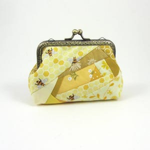Può includere: Un piccolo portamonete in tessuto con un design patchwork nei toni del giallo, crema e oro. Il tessuto presenta motivi a nido d'ape, piccoli fiori bianchi e api. È dotato di una chiusura a scatto in metallo color bronzo scuro.