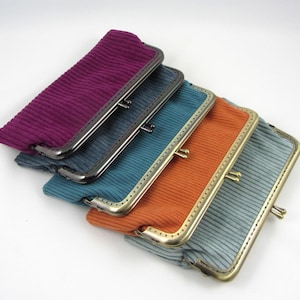Può includere: Una collezione di cinque portamonete in velluto a coste in vari colori: viola, verde acqua, arancione e grigio. Ogni portamonete ha una forma rettangolare con chiusura a scatto in metallo. I portamonete sono disposti in diagonale, mostrando i loro diversi colori e texture.
