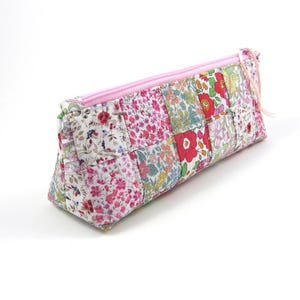 Peut inclure: Une trousse rectangulaire composée d'un patchwork de tissus à motifs floraux dans des tons de rose, rouge, vert et blanc. La trousse présente une fermeture éclair rose clair sur le bord supérieur avec une tirette en tissu assortie.