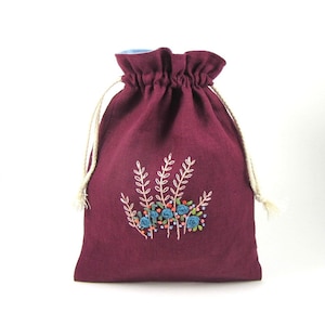 Könnte beinhalten: Eine burgunderfarbene Leinentasche mit Kordelzug und einem floralen Stickmuster mit blauen, rosa und grünen Blumen und cremefarbenen Stielen.