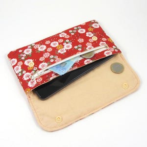 May include: Red floral fabric clutch with a zipper closure, open to reveal a smartphone, cash, and coins. The interior is beige, and the clutch is secured with snap closures. The floral pattern includes white and yellow flowers.