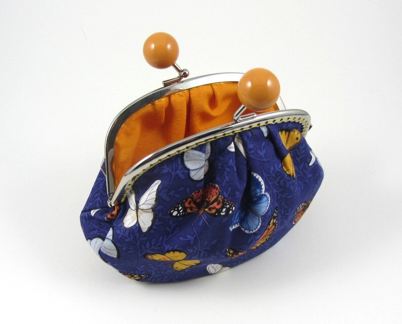 May include: A blue coin purse with a butterfly print and a silver clasp. The purse is open and the inside is lined with orange fabric.