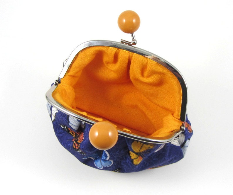 May include: A small blue coin purse with a butterfly print and a yellow snap closure. The purse is open and the inside is lined with orange fabric.