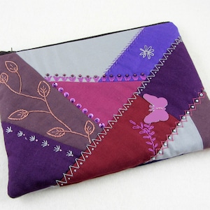 Op de afbeelding: Paarse, grijze en bordeauxrode patchwork stoffen ritszak met geborduurde bloemen en een vlinder.