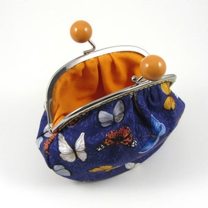 May include: A small blue coin purse with a butterfly print and a yellow trim. The purse is open and the inside is lined with orange fabric. The purse has two orange plastic clasps.