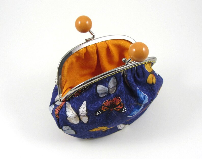 May include: A small blue coin purse with a butterfly print and a yellow trim. The purse is open and the inside is lined with orange fabric. The purse has two orange plastic clasps.