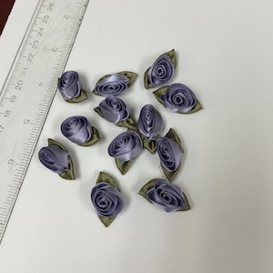 May include: A collection of 12 small, purple satin ribbon roses with green leaves. The roses are arranged on a white surface.