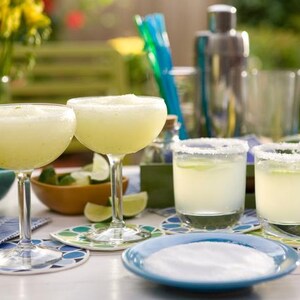 Puede incluir: Dos c&oacute;cteles de margarita en copas de tallo y dos en vasos bajos, todos con una rodaja de lima como adorno. Tambi&eacute;n se ve un plato azul con sal, limas y una coctelera. Las bebidas est&aacute;n sobre posavasos estampados.