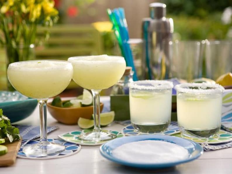 Puede incluir: Dos c&oacute;cteles de margarita en copas de tallo y dos en vasos bajos, todos con una rodaja de lima como adorno. Tambi&eacute;n se ve un plato azul con sal, limas y una coctelera. Las bebidas est&aacute;n sobre posavasos estampados.