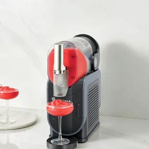 Puede incluir: Una m&aacute;quina de granizados negra y gris con una parte superior transparente, que dispensa una bebida roja en una copa con pie. Una botella vierte el l&iacute;quido rojo en la m&aacute;quina. Una bebida terminada est&aacute; en el mostrador.