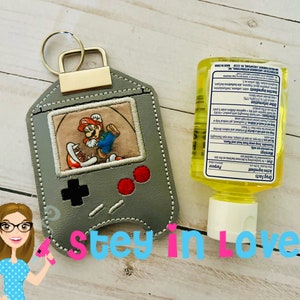 May include: A gray and black Game Boy-style keychain with a picture of Super Mario in the screen. The keychain has a gold ring and is attached to a small bottle of hand sanitizer.