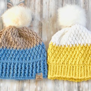 May include: Two handmade crochet beanies with faux fur pom-poms. One is blue and beige, the other is white and yellow. Both have a textured stitch pattern and a tag that reads "Peanut & Plum".