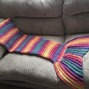 May include: A handmade, rainbow-colored crochet mermaid tail blanket. The blanket features a striped pattern with shades of yellow, orange, pink, purple, blue, and green. The tail is fanned out at the end. The blanket is laying on a gray couch.