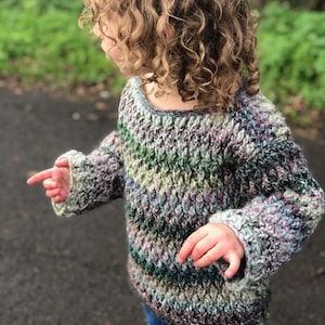 May include: A child wearing a green, purple and blue striped crocheted sweater with long sleeves. The sweater has a loose fit and a slightly dropped shoulder.