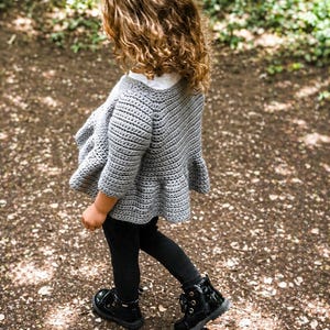 May include: A gray, crocheted cardigan with a ruffled hem. The child is wearing black leggings and shiny black boots with laces. The background is a blurred outdoor scene with sunlight.