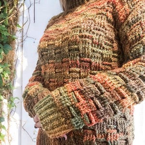 Op de afbeelding: Een handgebreide trui met een mandweefpatroon. De trui heeft een hoge hals en lange mouwen. Het kleurenpalet omvat tinten bruin, oranje en groen. De trui is gemaakt van dik garen, waardoor hij een textuur heeft.