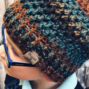 May include: A handmade, textured beanie in shades of brown, teal, and orange. The hat features a woven pattern and a small tag that reads "Peanut & Plum". The beanie is worn with blue-framed glasses.