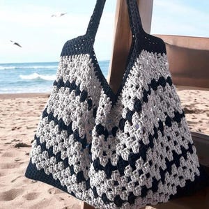 Op de afbeelding: Een handgemaakte gehaakte tote bag in een geometrisch patroon van grijs en marineblauw. De tas heeft lange hengsels en wordt getoond aan een houten stoel op het strand. Het open geweven ontwerp van de tas is zichtbaar.