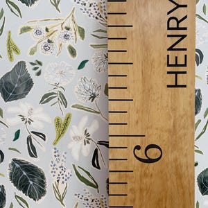 May include: A wooden growth chart with a light brown stain and black markings. The chart has a black inscription that reads "HENRY" and a black number "6" at the bottom. The chart is set against a light blue background with a floral pattern.