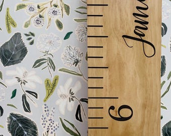 May include: Wooden growth chart with the name "James" in cursive script. The chart has black measurement markings and the number 6. The background is a light blue floral pattern with white and green leaves and flowers.