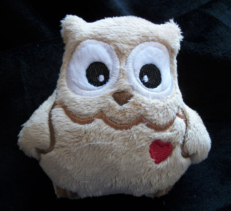May include: A plush owl toy with embroidered eyes, a brown beak, and a red heart on its chest. The owl is beige and has a soft, fuzzy texture.