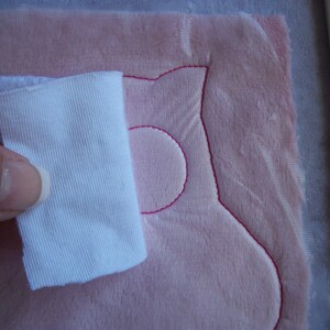 May include: A white fabric scrap is being used to smooth out a pink fabric with a pink embroidered design. The design is a simple, rounded shape with a circle in the middle.