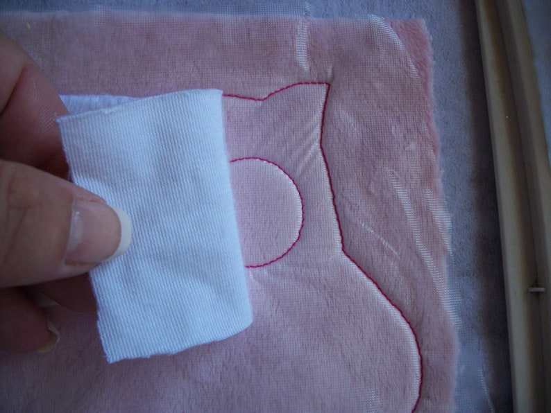 May include: A white fabric scrap is being used to smooth out a pink fabric with a pink embroidered design. The design is a simple, rounded shape with a circle in the middle.