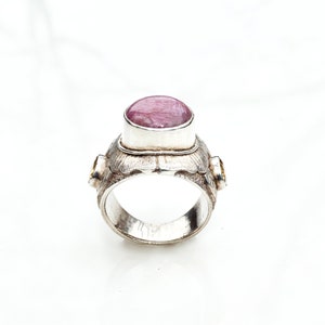 Può includere: Anello d'argento con una grande pietra preziosa rosa ovale. L'anello ha una fascia strutturata e due pietre preziose gialle più piccole. L'anello è su sfondo bianco. Un gioiello.