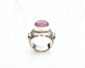 May include: A silver ring with a large, oval, pink gemstone as the centerpiece. The ring has a textured band and two smaller yellow gemstones on either side of the main stone. The ring is set against a white background.