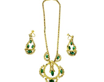 May include: A gold necklace and earrings set with a green rhinestone design. The necklace features a large pendant with a circular design and the earrings are teardrop shaped.