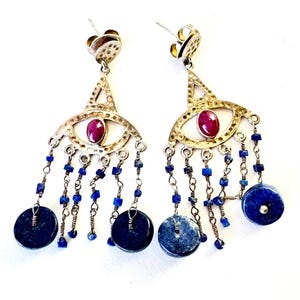 May include: A pair of silver-coloured drop earrings. Each earring has an eye-shaped design with a red gemstone centre, dangling chains with blue beads, and a large, dark blue circular stone at the bottom. The earrings have a post-back closure.