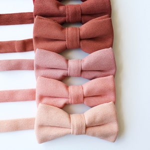 May include: A collection of five bow ties in varying shades of red and pink, arranged vertically. The bow ties are made of a textured fabric, with matching adjustable neckbands. The colors range from deep burgundy to light peach.