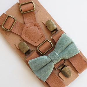 May include: A set of brown leather suspenders with antique brass-colored hardware and a light green bow tie. The suspenders are displayed on a brown cardboard backing.