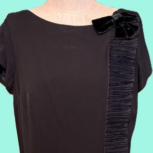 May include: A black dress with short sleeves and a straight silhouette. A vertical strip of pleated fabric runs down the right side. A black velvet bow is at the shoulder. The dress is displayed on a mannequin.