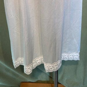 Op de afbeelding: Lichtblauwe onderrokrok op een mannequin. De rok heeft een witte elastische tailleband en een delicate witte kanten rand aan de zoom. De stof is een lichtgewicht, semi-transparant materiaal.