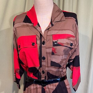Peut inclure: Une robe à motifs rouge, noir et marron avec un devant boutonné et une ceinture noire. La robe a un col et des manches courtes.