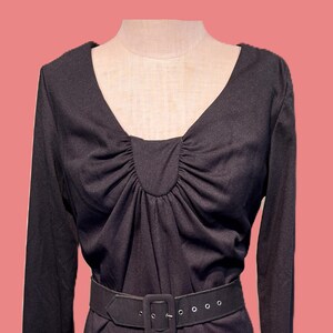 May include: A long-sleeved black dress featuring a V-neckline with gathered fabric detailing at the center chest. The dress includes a matching black belt with a rectangular buckle and silver-toned eyelets, cinched at the waist for a structured fit.