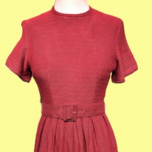 May include: A red vintage dress with short sleeves and a fitted waist. The dress features a textured fabric with diagonal stripes and a matching belt with a square buckle. The skirt is pleated, and the dress has a round neckline.